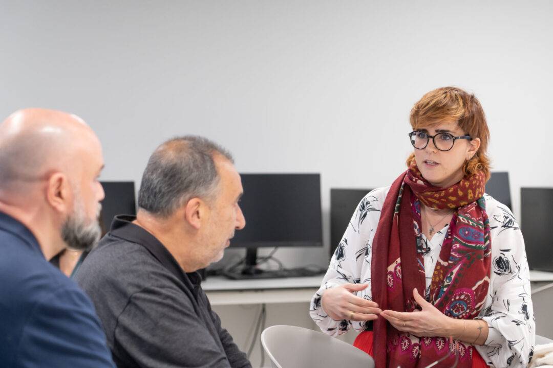 Una mujer habla mientras dos hombres la escuchan en un contexto de reunión.
