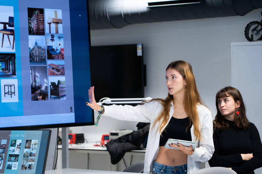 Una joven presenta un proyecto en una pantalla mientras otra persona la observa.