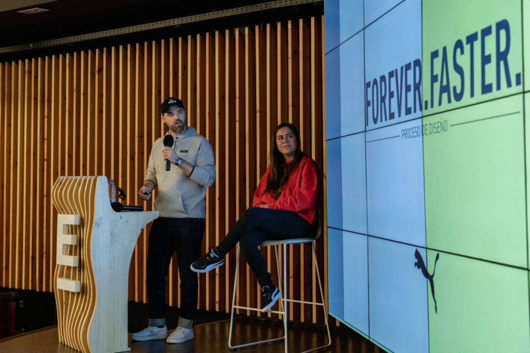Dos personas participan en una charla en un evento, con una pantalla detrás que muestra el lema 'FOREVER.FASTER.'
