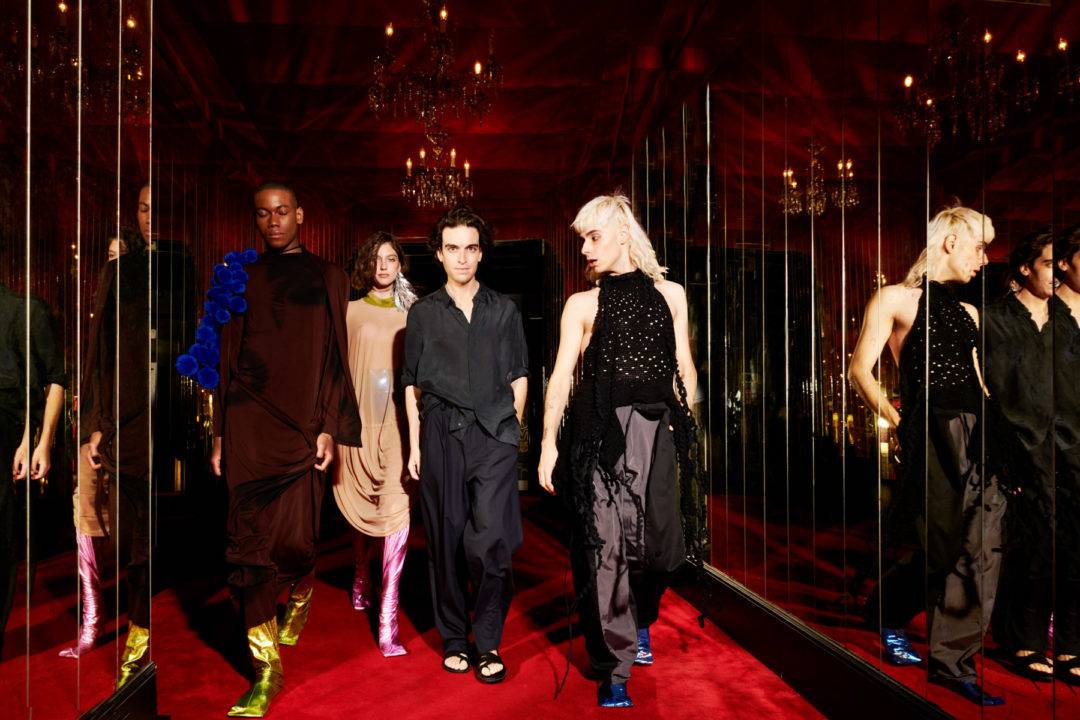 Un grupo de modelos camina por un pasillo con espejos en un ambiente elegante y colorido.