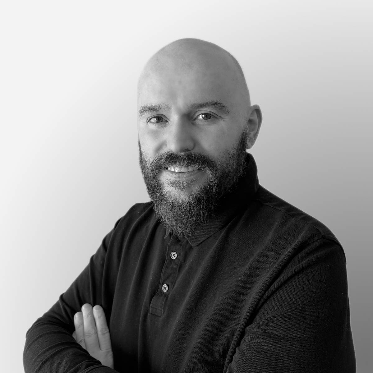 Retrato en blanco y negro de un hombre con barba y cabeza rapada, sonriendo y cruzando los brazos.