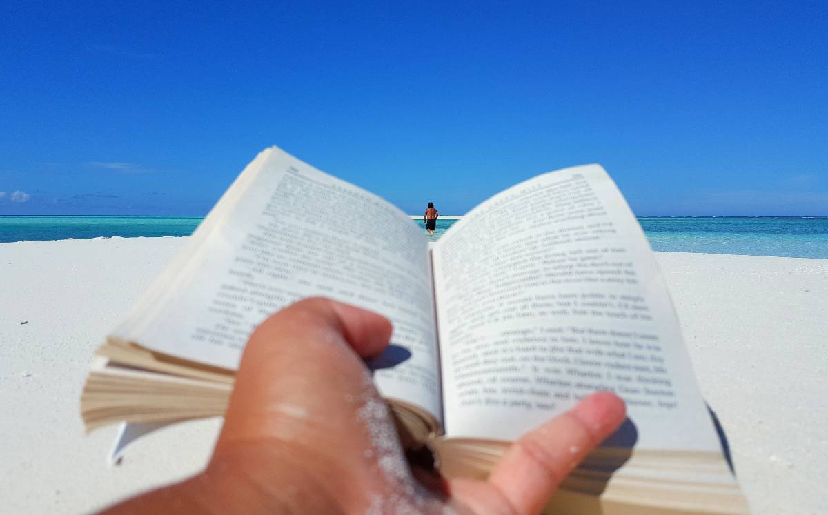 Una persona leyendo un libro en una playa con arena blanca y agua clara.