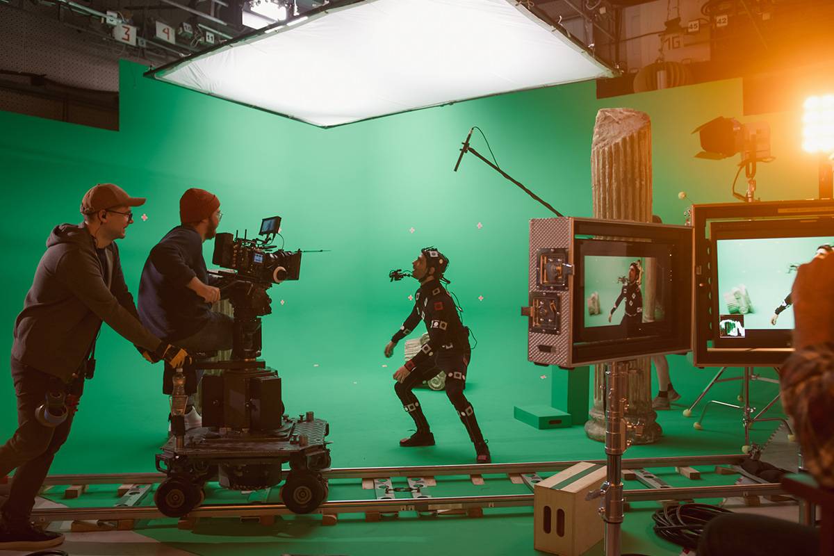 Una escena de grabación en un plató con fondo verde, donde un actor utiliza un traje de captura de movimiento mientras un camarógrafo lo filma.