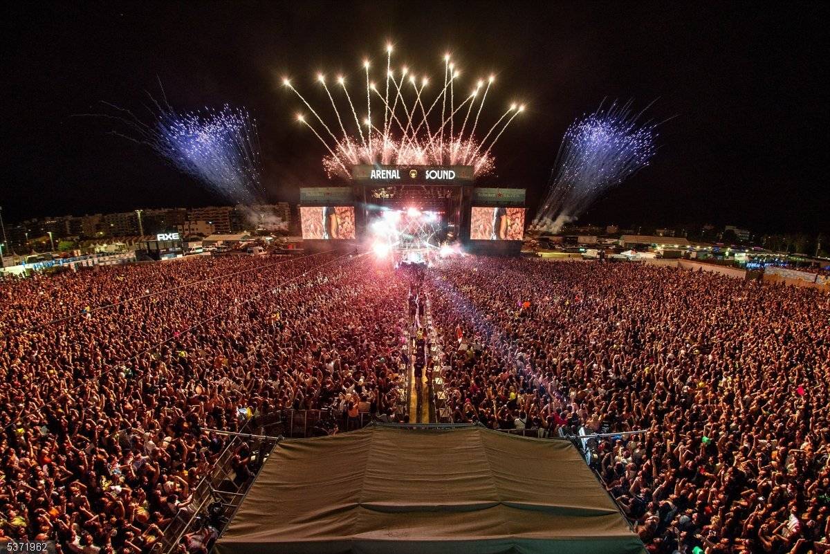 La imagen muestra un gran concierto al aire libre con un público numeroso y fuegos artificiales en el fondo.