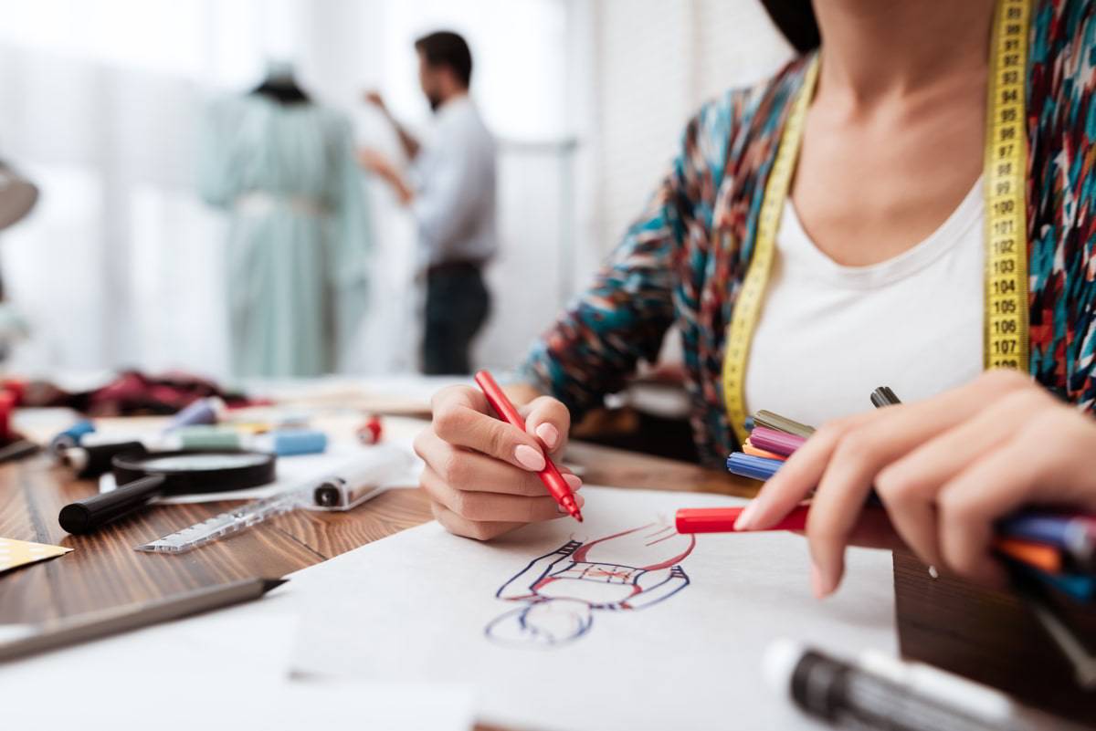 Una persona dibujando un diseño de moda mientras otra persona trabaja en el fondo.