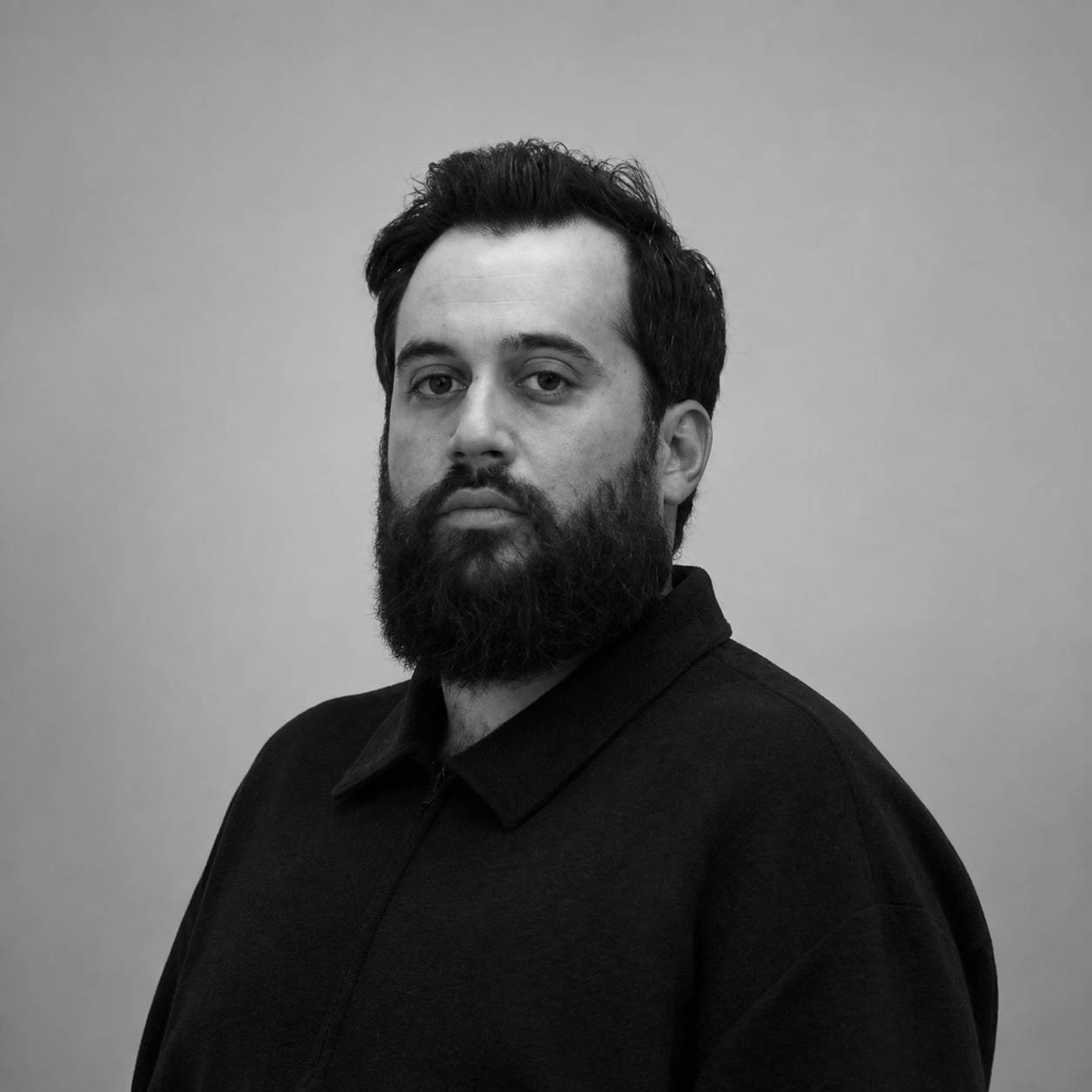 Un hombre con barba y cabello oscuro mira a la cámara, vestido con una chaqueta negra sobre un fondo gris.