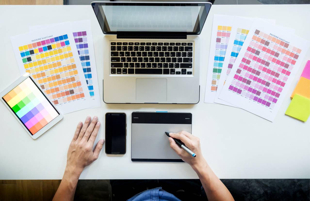 Una persona trabaja en una mesa con una computadora portátil, una tableta gráfica y varias muestras de color en papel.