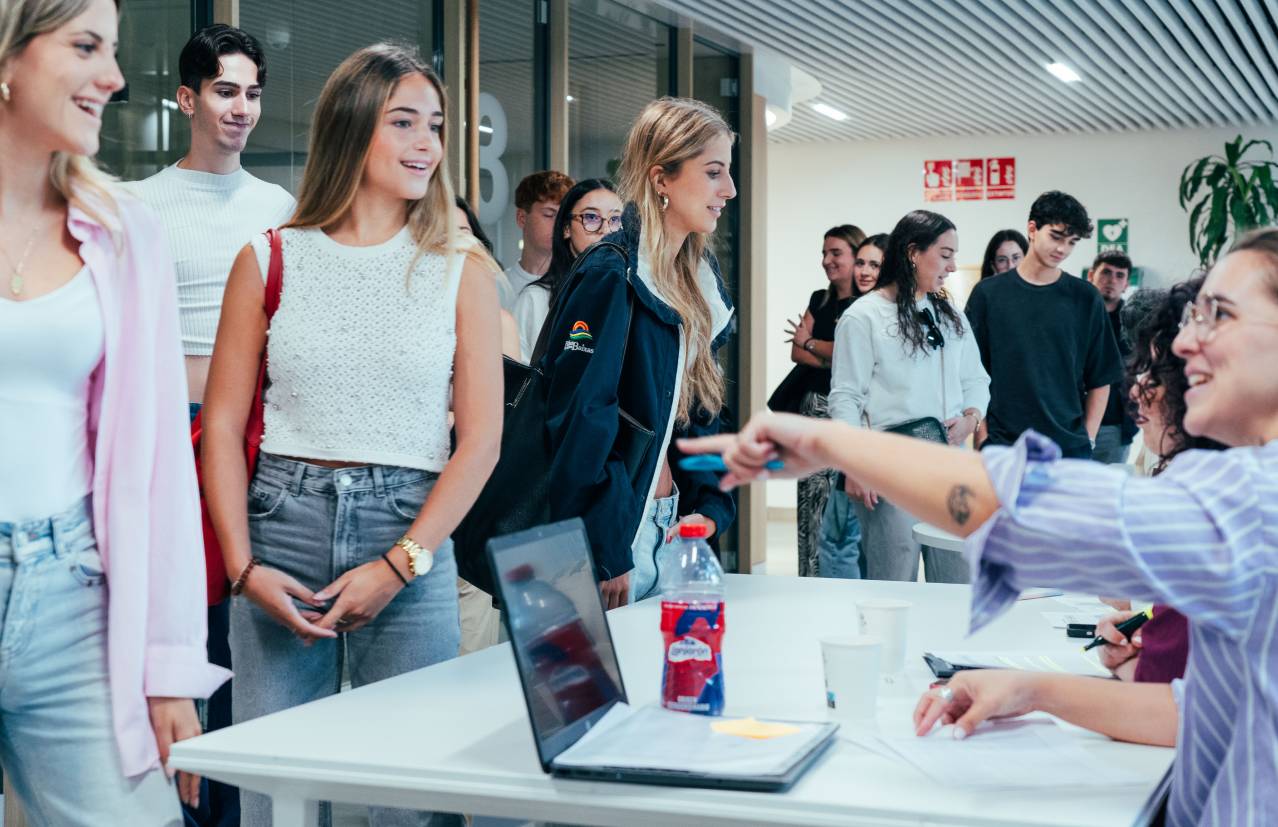 Un grupo de personas en un ambiente social donde se ven estudiantes interactuando y una persona en una mesa asistiendo a ellos.