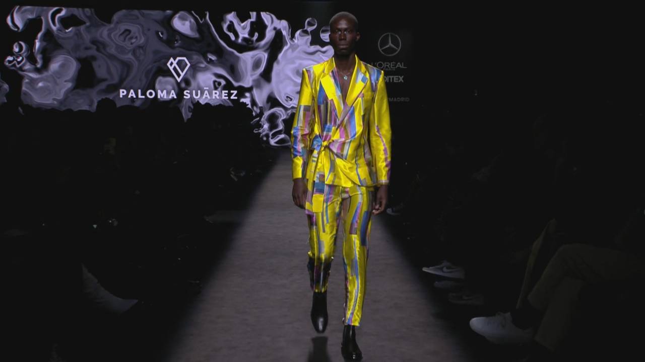 Un modelo camina por la pasarela luciendo un traje amarillo con patrones coloridos en un desfile de moda.