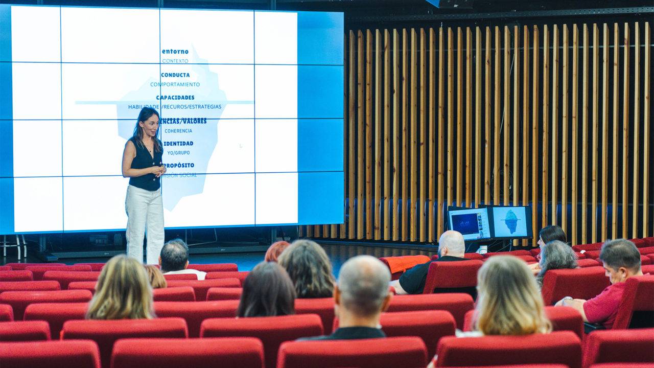 Una presentadora expone en un auditorio frente a un público atento mientras se proyectan diapositivas en una pantalla grande.