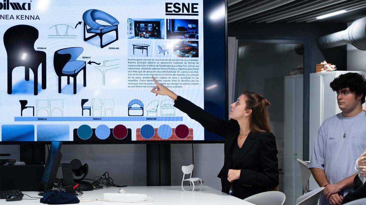 Una mujer explica un diseño de muebles frente a una pantalla en una sala de clases.