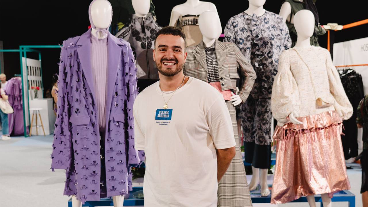 Un hombre sonriente posa frente a maniquíes que muestran diversas prendas de moda.