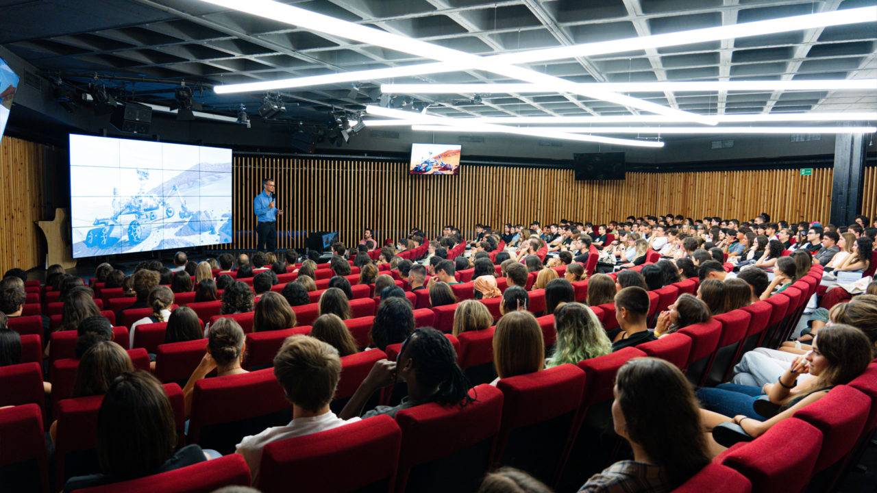 Una sala de conferencias llena de jóvenes escuchando a un ponente frente a una pantalla.