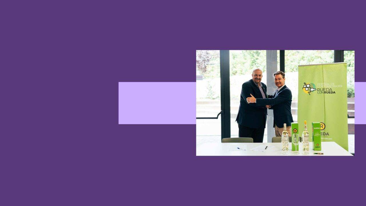 Dos hombres sonrientes se estrechan la mano ante una mesa con botellas y una bandera de fondo.