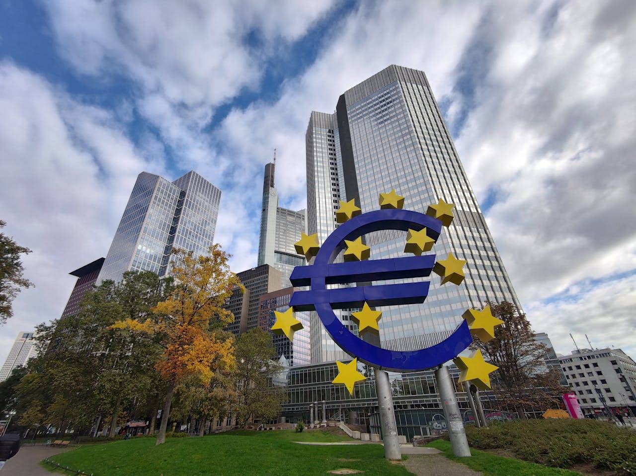 Escultura del símbolo del euro rodeada de edificios modernos y árboles en un entorno urbano.