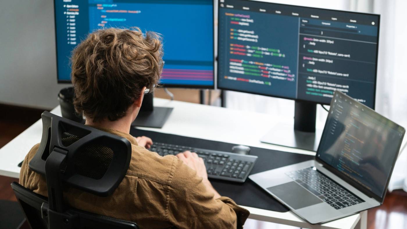 Una persona trabaja en su computadora portátil mientras codifica en dos monitores grandes.