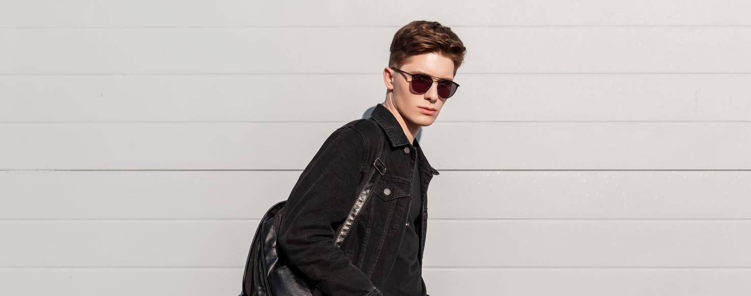 Un joven vestido de negro con gafas de sol y una mochila camina frente a una pared clara.