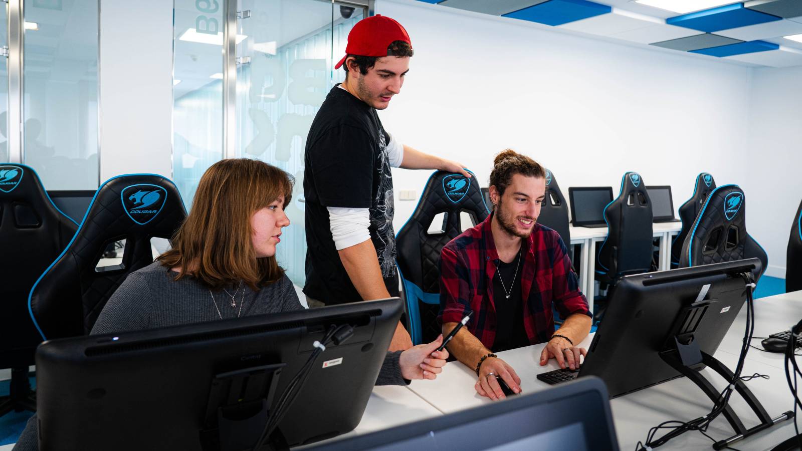 Un grupo de tres jóvenes colabora frente a unos ordenadores en un ambiente de trabajo moderno.
