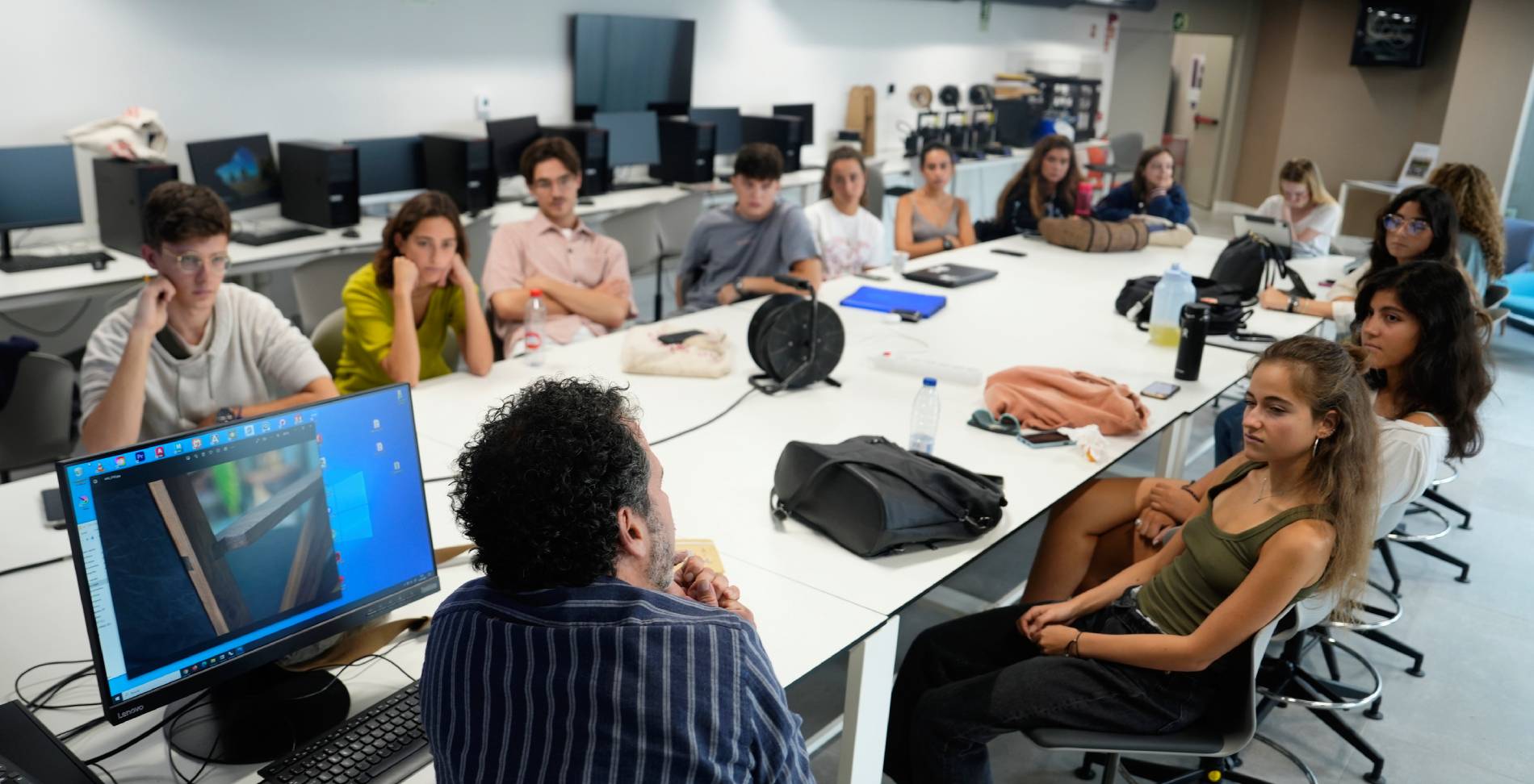 Un grupo de estudiantes escucha atentamente a un profesor durante una clase en un aula con computadoras.