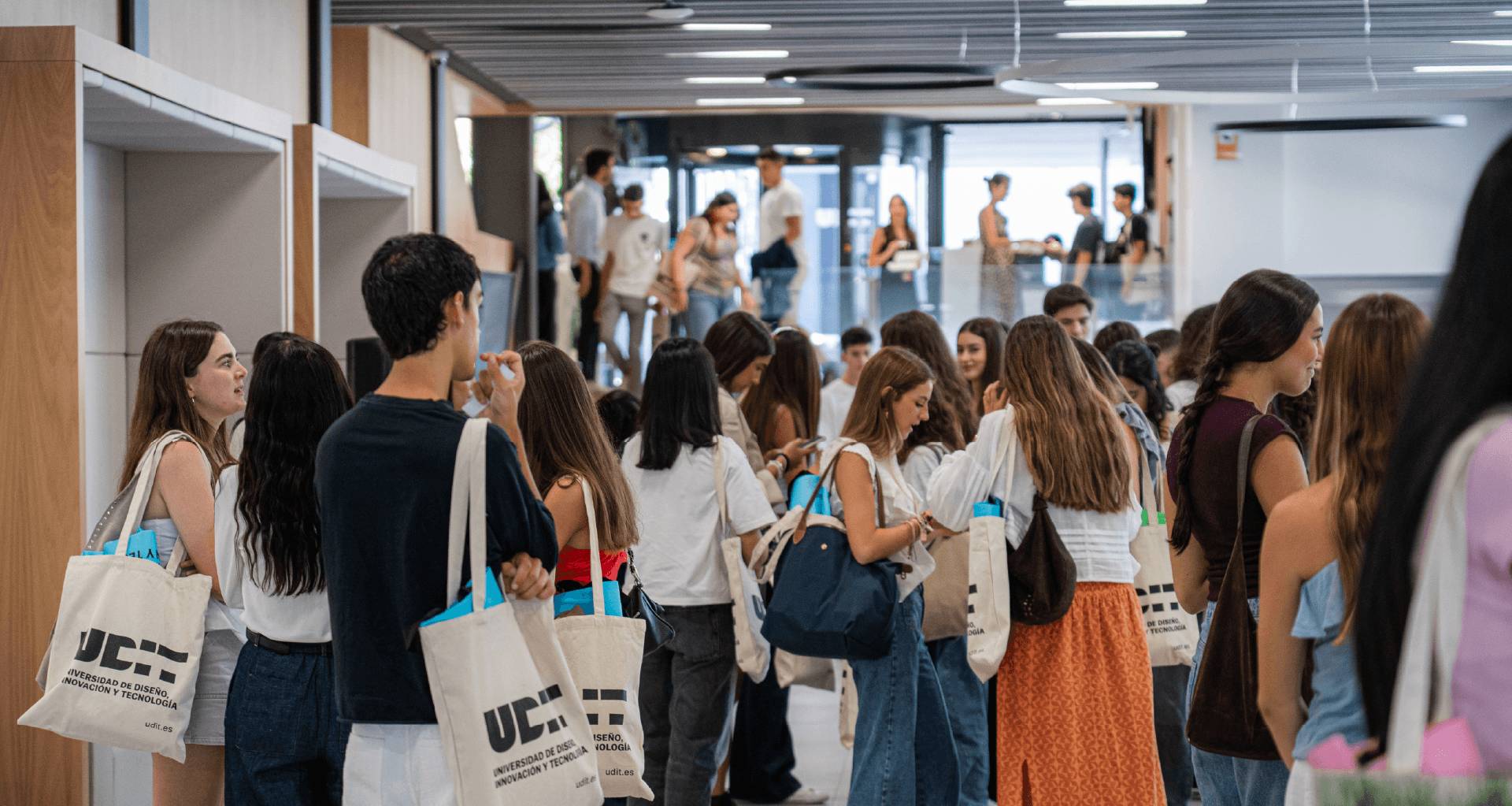 Un grupo de estudiantes se encuentra en un edificio moderno, algunos de ellos llevan bolsas con el logo de la universidad.