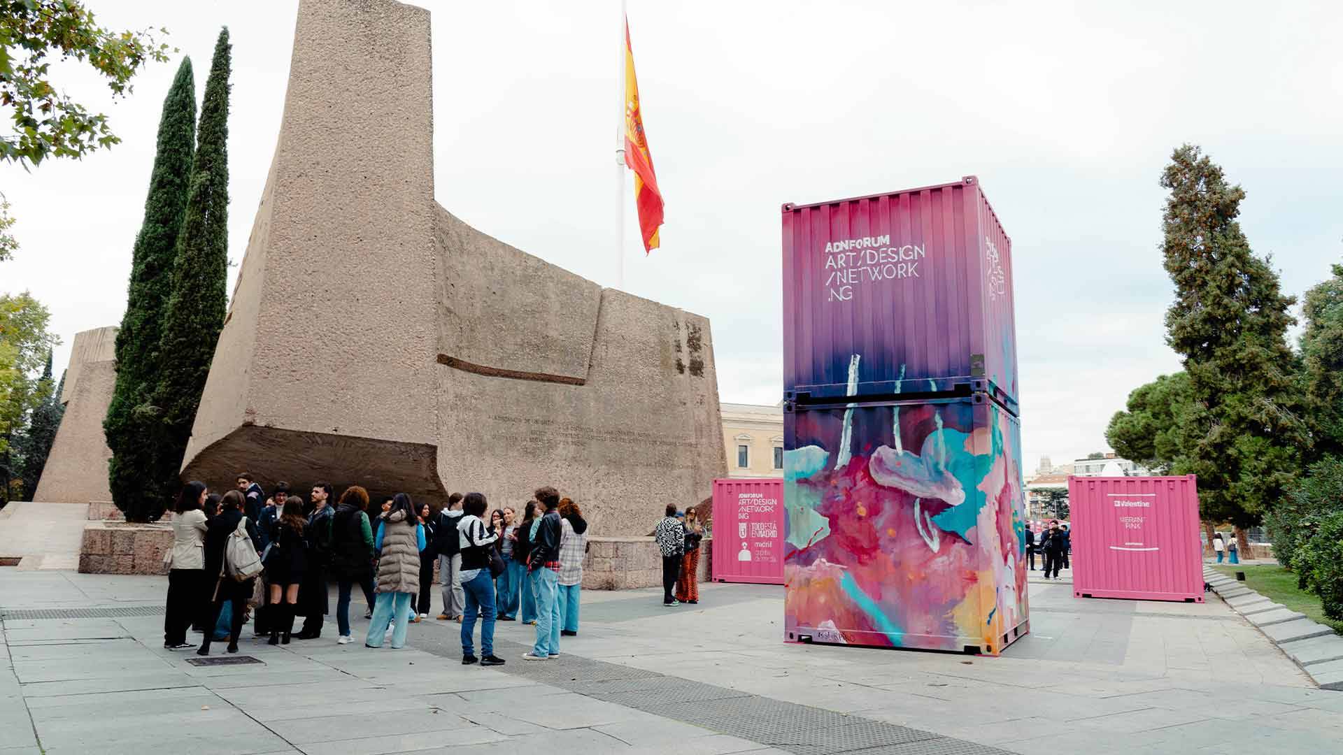 En la imagen se observa un grupo de personas frente a un contenedor artístico y una escultura monumental con una bandera ondeando en el fondo.