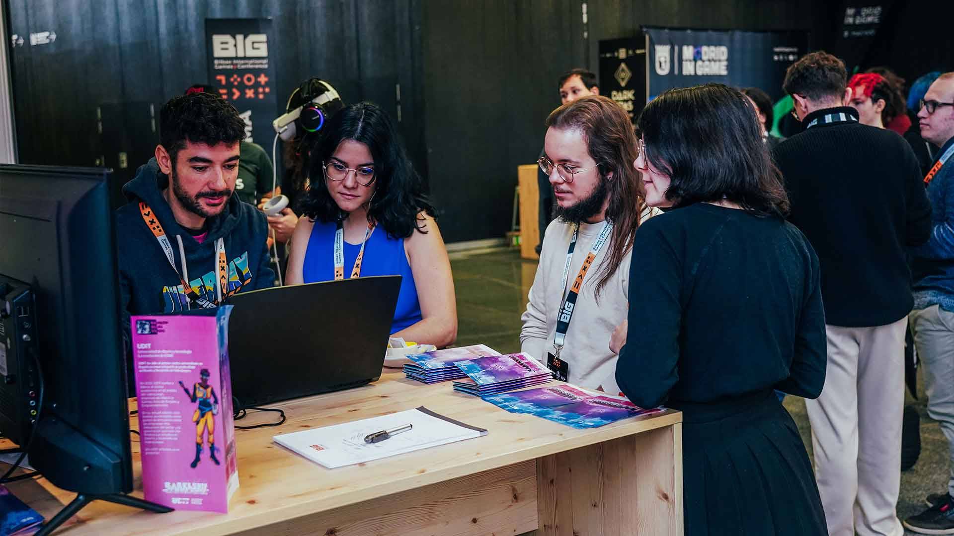 Un grupo de personas interactúa en un stand durante un evento, mirando una pantalla de computadora.