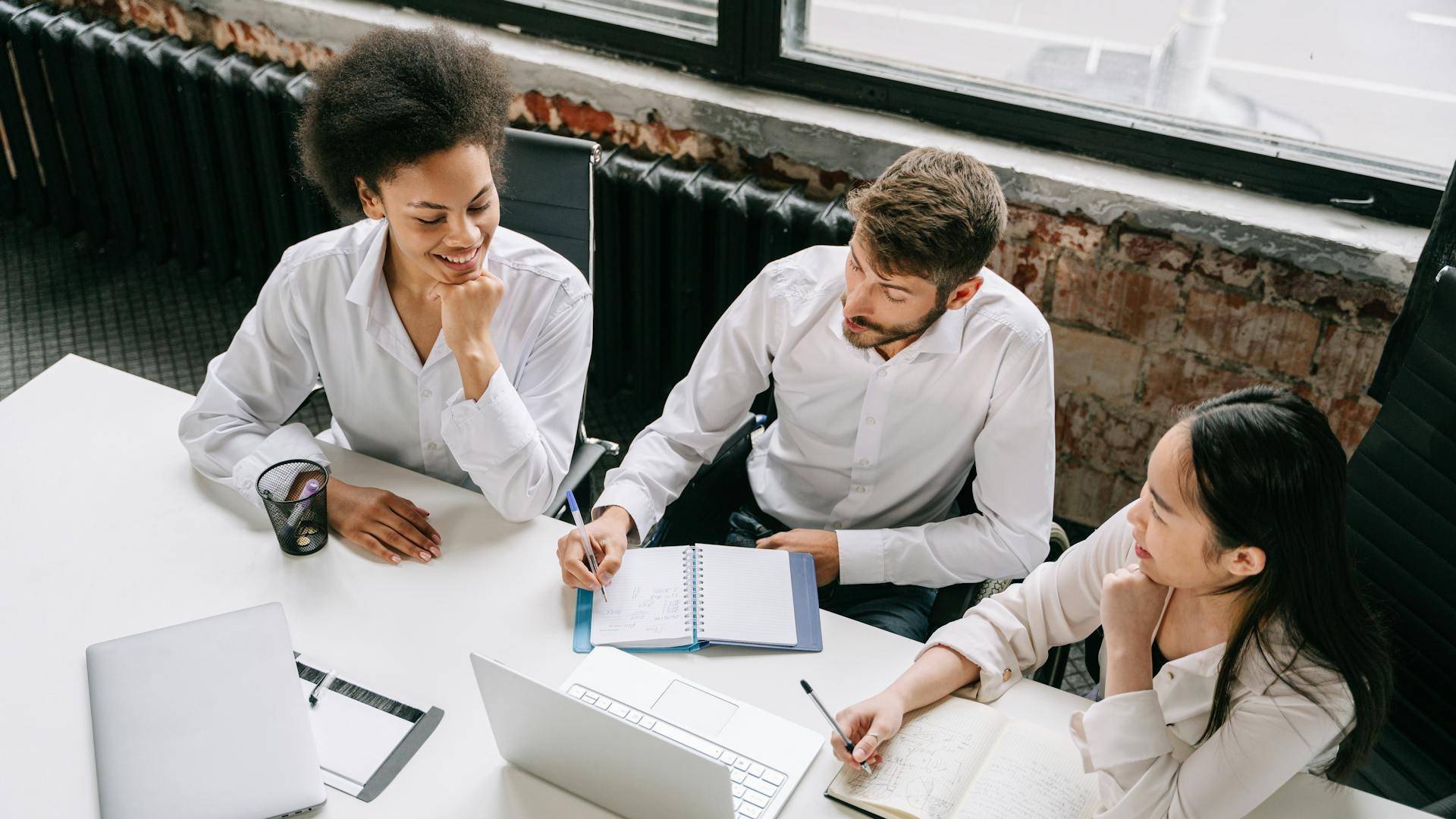 Un grupo de tres personas, dos mujeres y un hombre, están trabajando juntos en una mesa con computadoras portátiles y cuadernos.