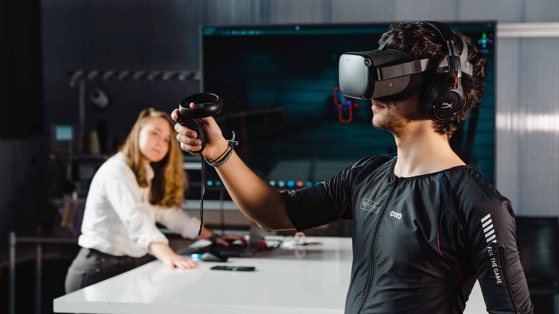 Un hombre está utilizando un visor de realidad virtual mientras sostiene un controlador, mientras una mujer lo observa desde el fondo.