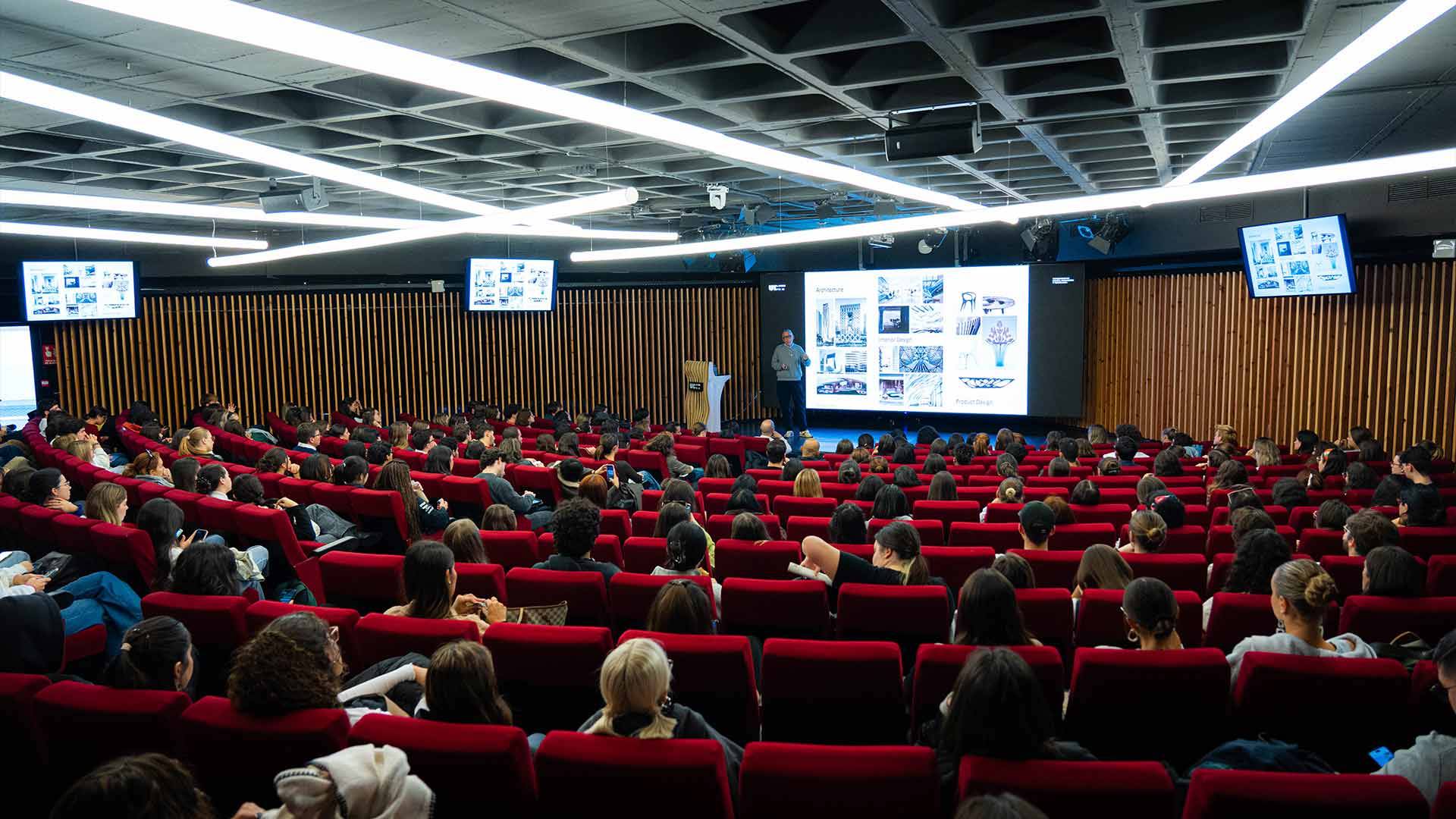 Un gran auditorio con una multitud de personas sentadas mientras un presentador expone en una pantalla.