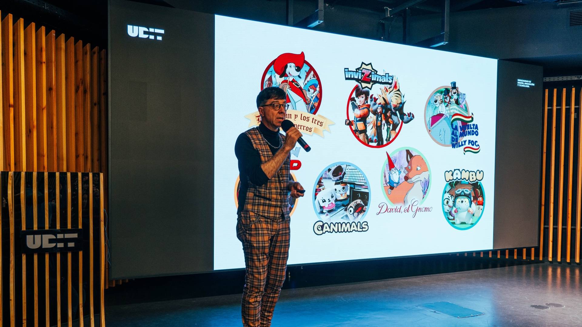 Un presentador habla frente a una pantalla que muestra una serie de ilustraciones y logotipos relacionados con diferentes personajes y temas.