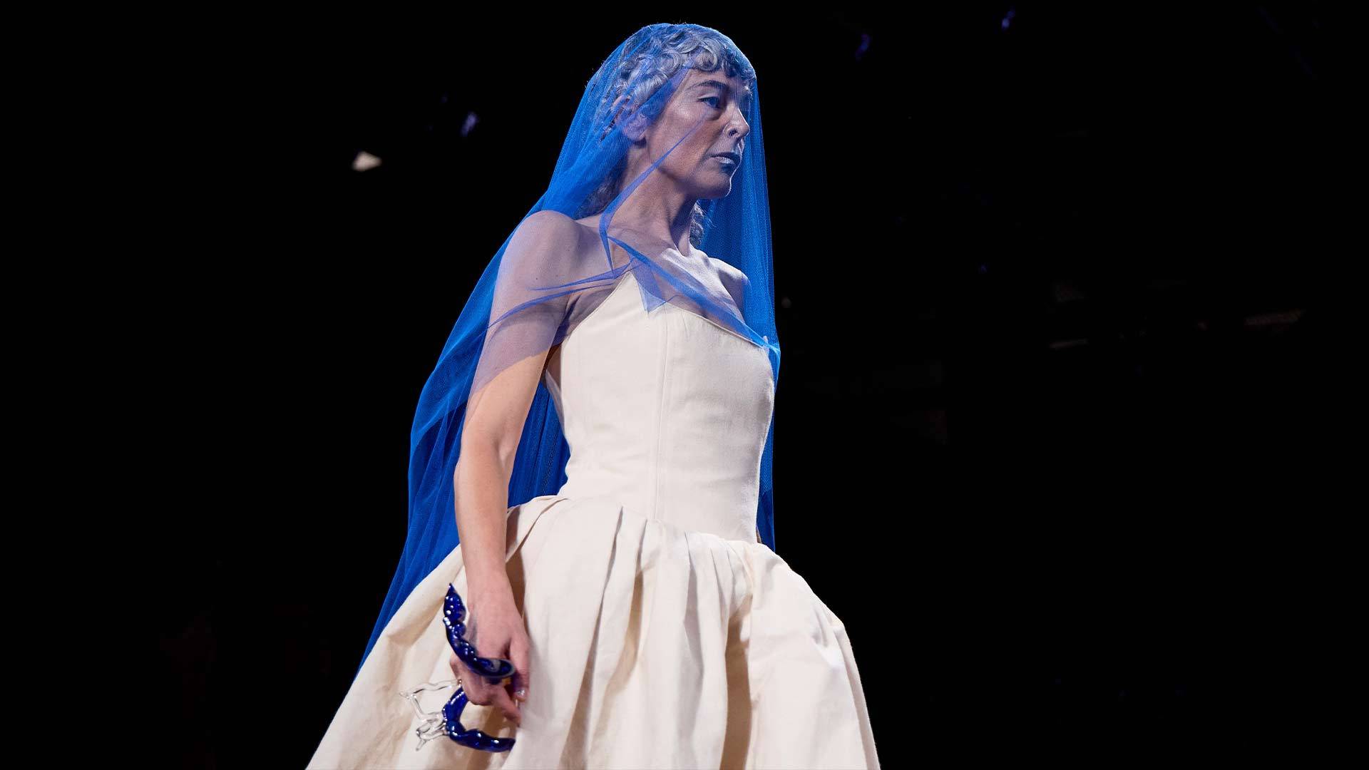 Una modelo presenta un vestido blanco con un velo azul en un desfile de moda.
