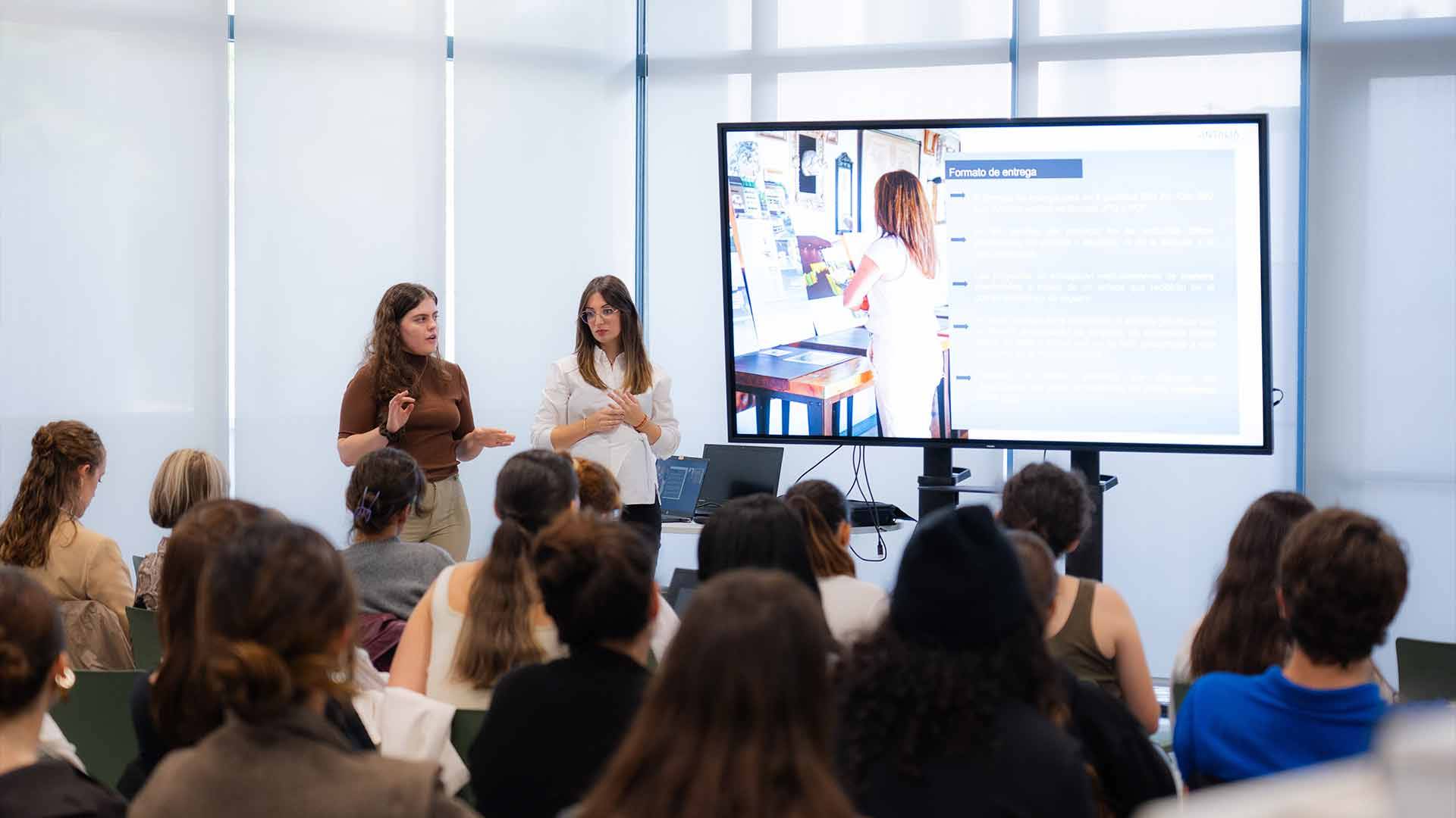 Una presentación en una sala con una audiencia atenta y dos ponentes frente a una pantalla.