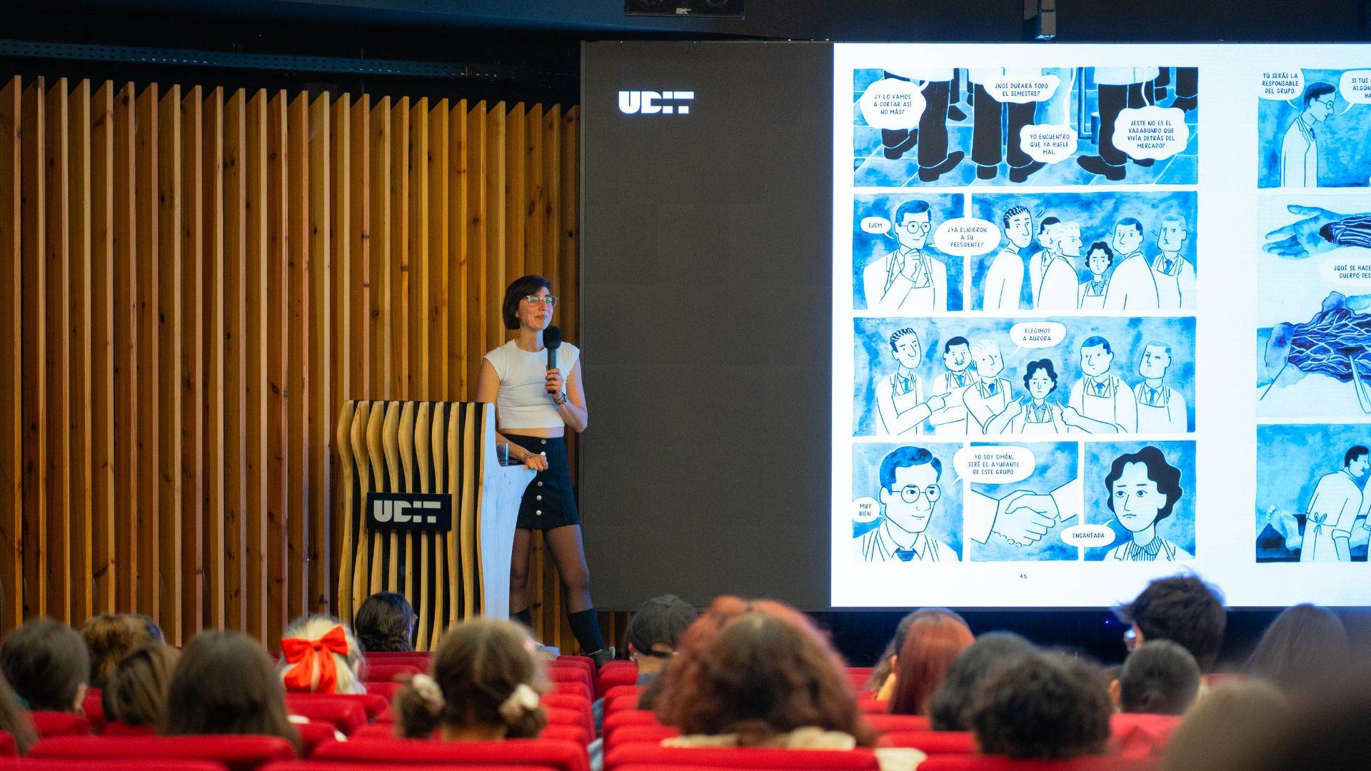 Una presentadora expone ante una audiencia en un auditorio mientras se proyectan imágenes en una pantalla grande.