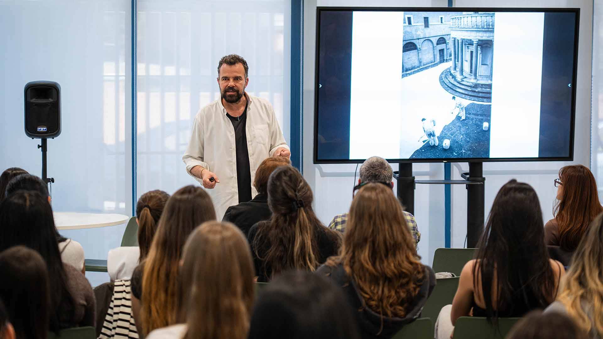 Un presentador está hablando frente a una audiencia mientras muestra una imagen en una pantalla.