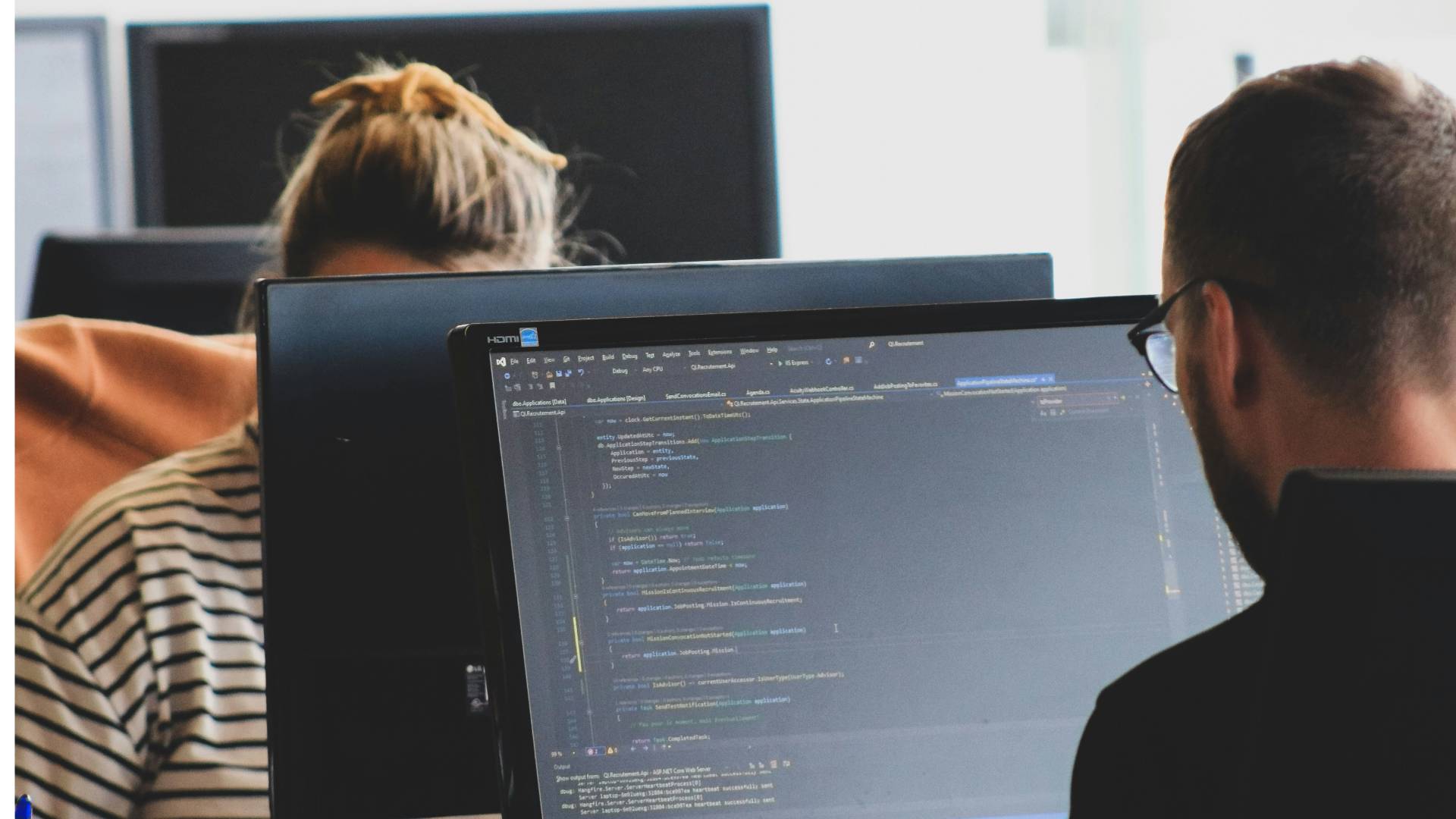 Dos personas trabajando en oficinas, una de espaldas frente a un ordenador, y otra con una camiseta de rayas parcialmente visible.