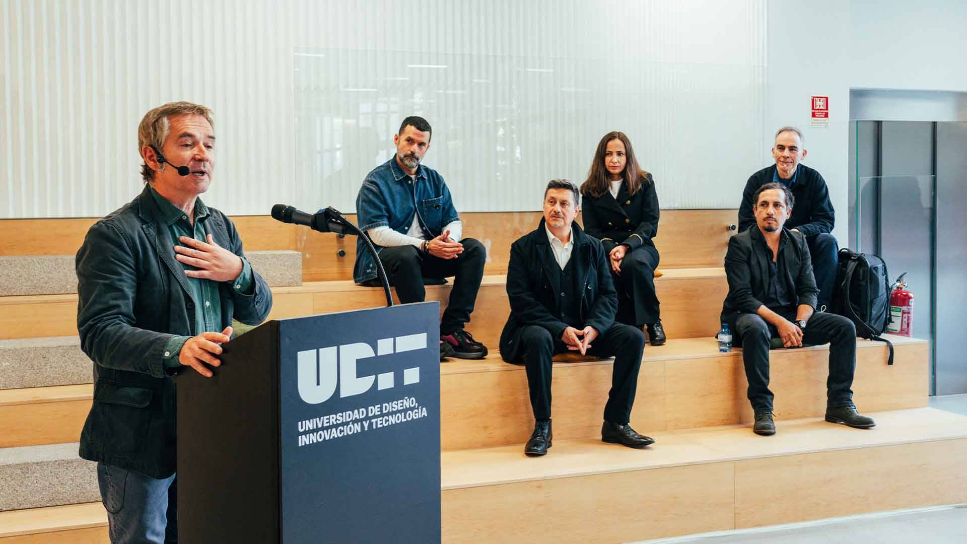 Un evento académico en el que varios ponentes están presentes en un escenario mientras uno está hablando frente a un micrófono.