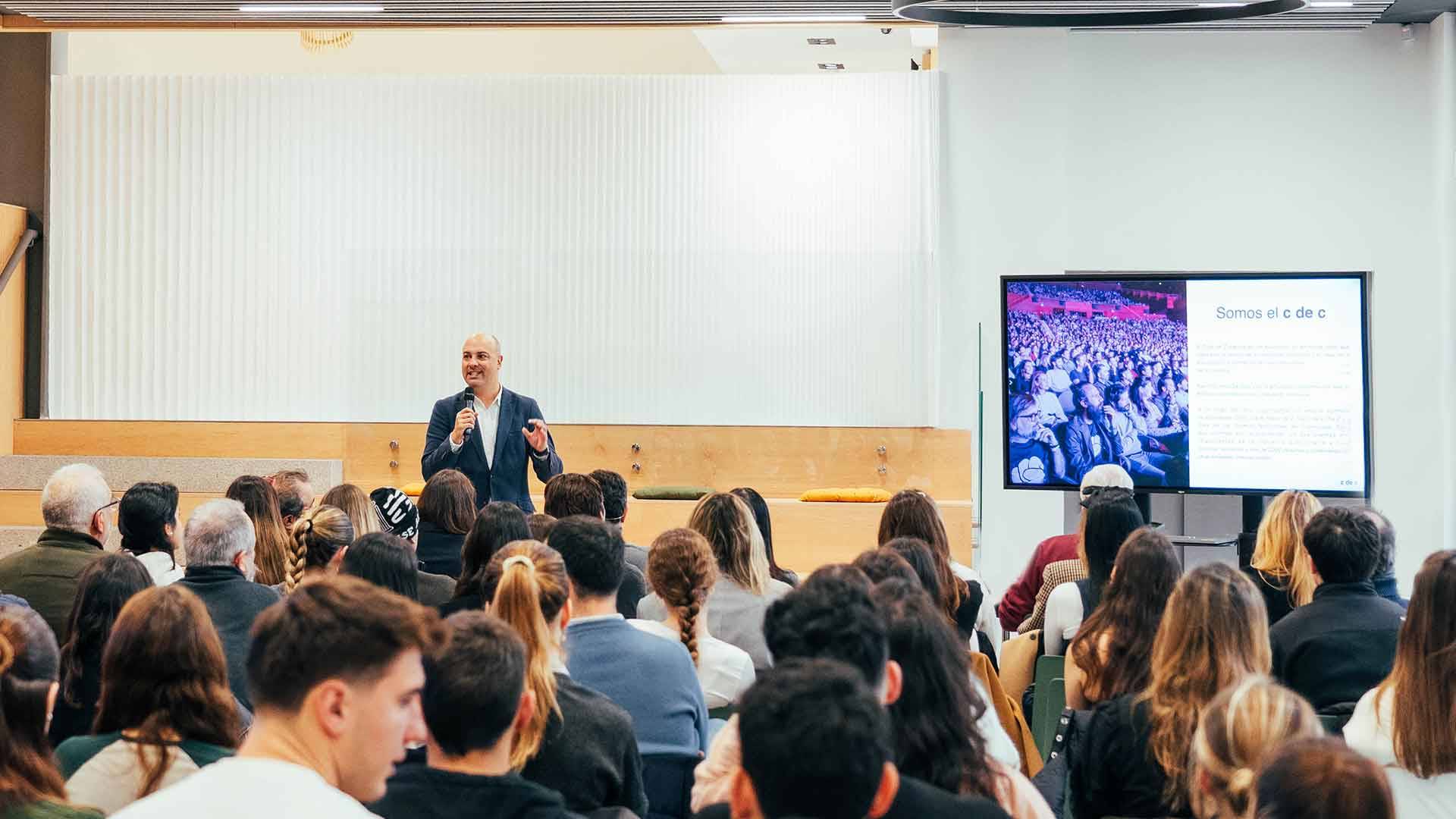 Un orador se presenta ante una audiencia en un evento, mientras una pantalla muestra información al fondo.