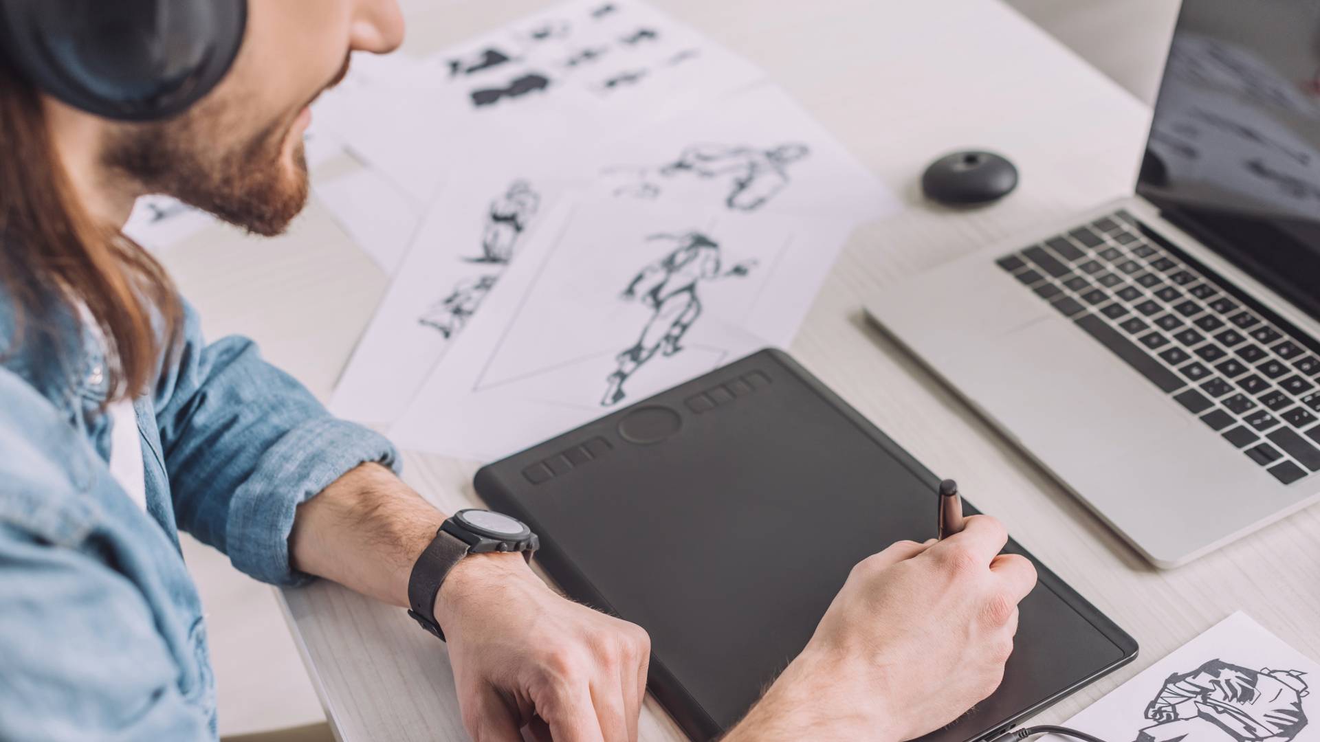 Una persona está dibujando en una tableta gráfica mientras escucha música y observa hojas de papel con bocetos.