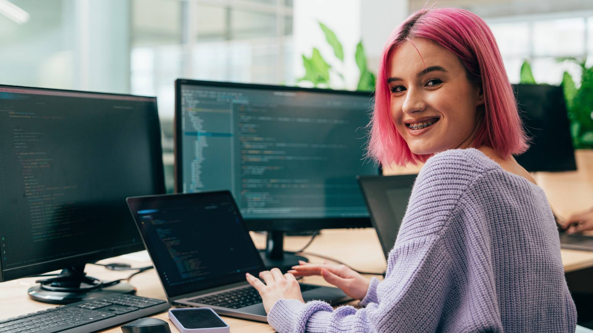 Una joven con cabello rosa sonríe mientras trabaja en su laptop rodeada de pantallas de computadora.