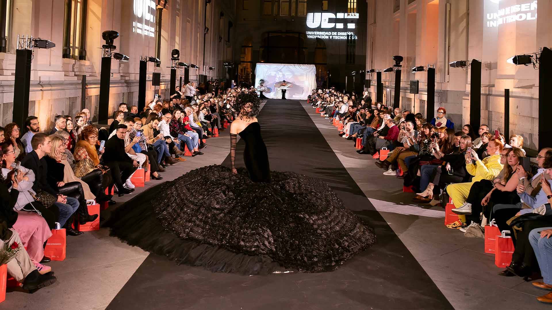 Una pasarela de moda con un modelo vistiendo un vestido negro de gran volumen ante un público aplaudiendo.