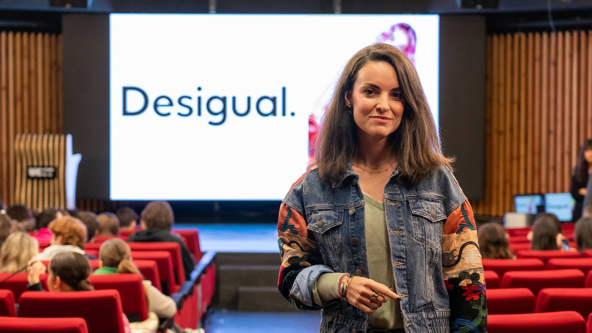 Una mujer sonriente presenta un evento sobre la marca Desigual en un auditorio con público.