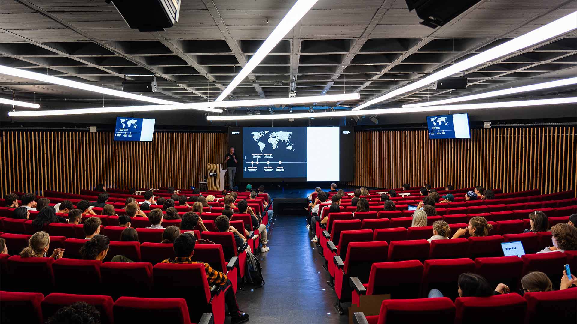 La imagen muestra a un presentador exponiendo en un auditorio lleno de personas sentadas.