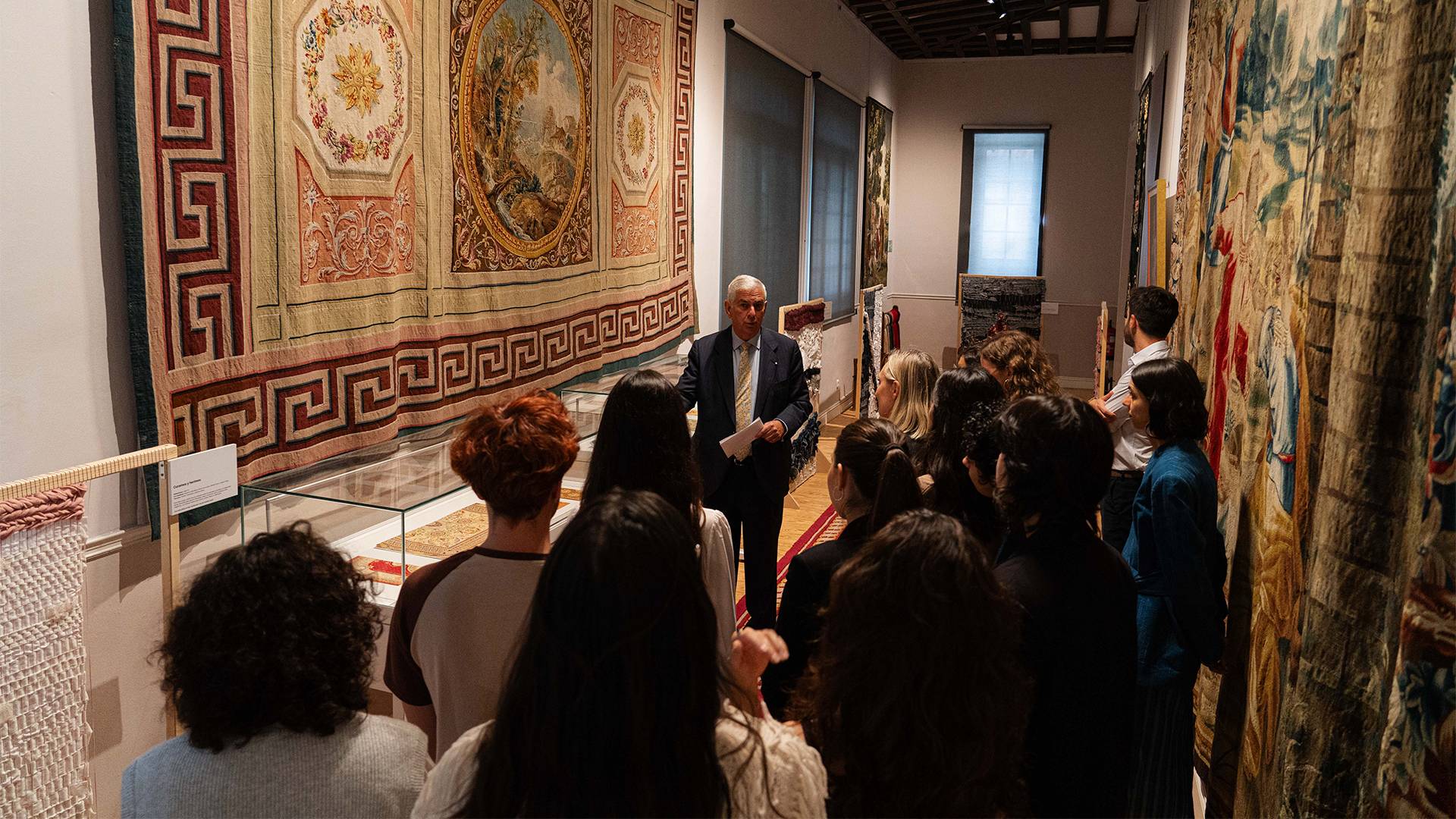 Una visita guiada en un museo con tapices en las paredes y un grupo de personas escuchando a un hablante.
