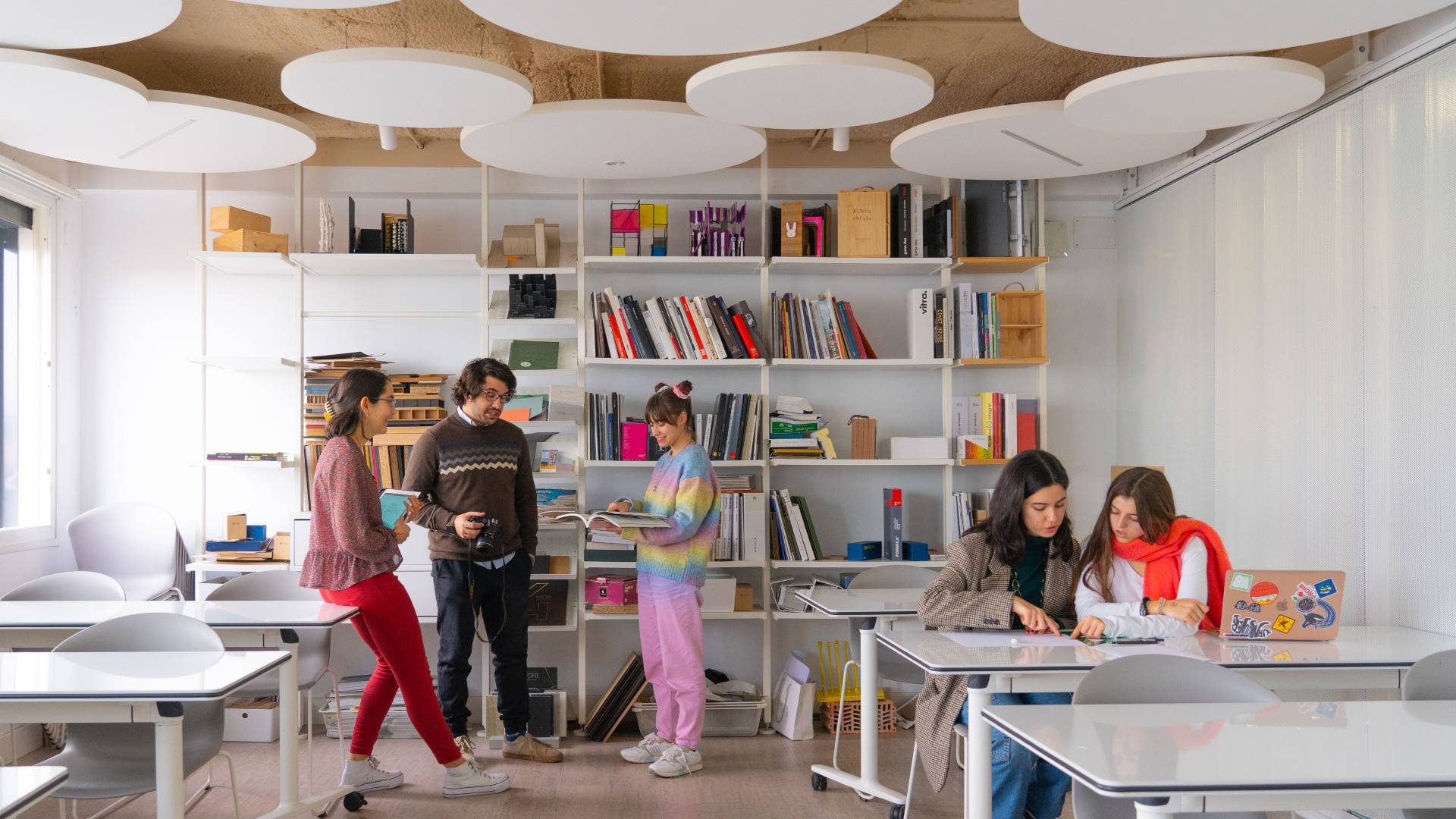 Un grupo de estudiantes interactúa en un aula moderna con estanterías llenas de libros y materiales de estudio.