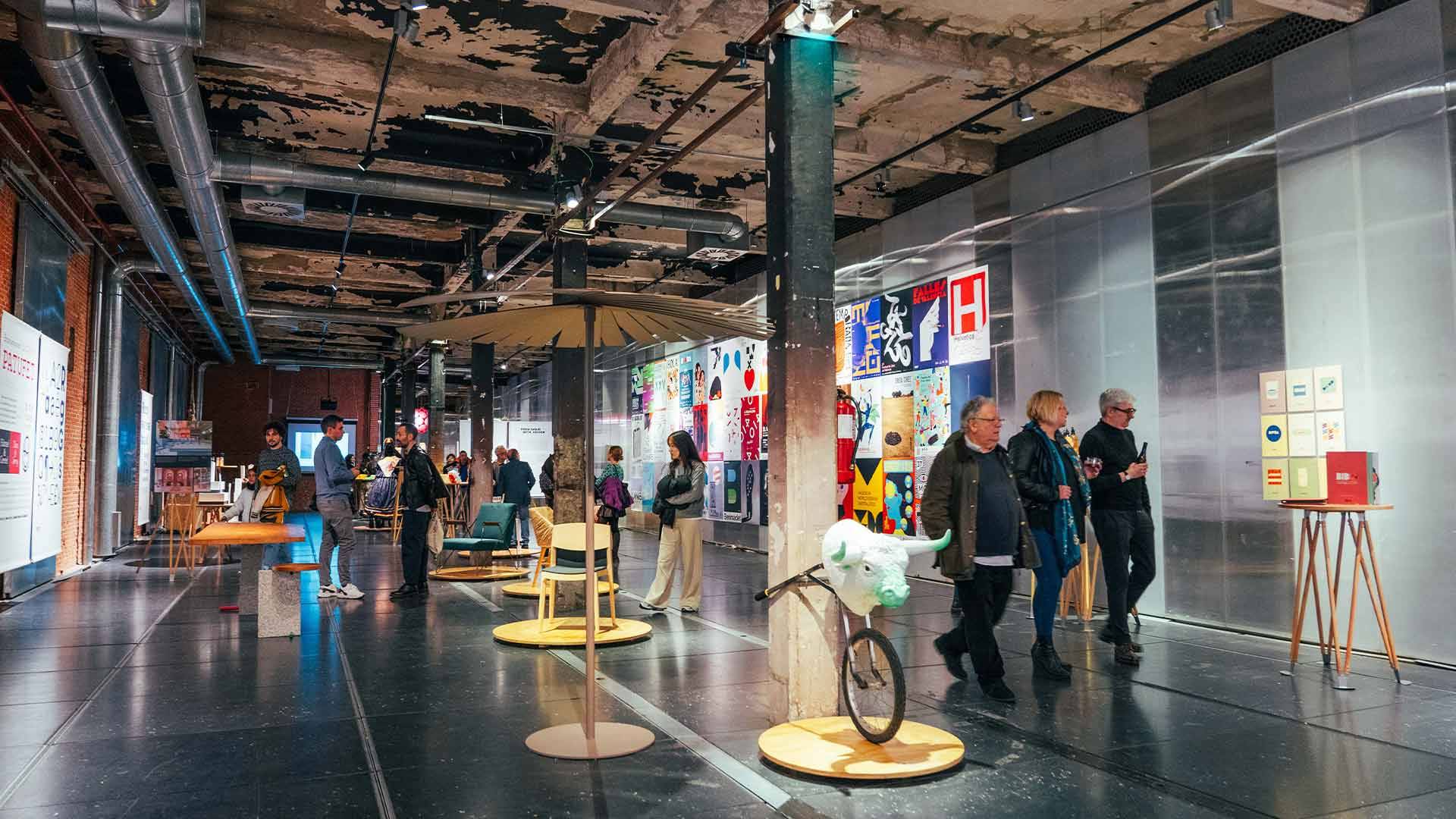 La imagen muestra una exposición artística en un espacio contemporáneo con personas interactuando con las obras.