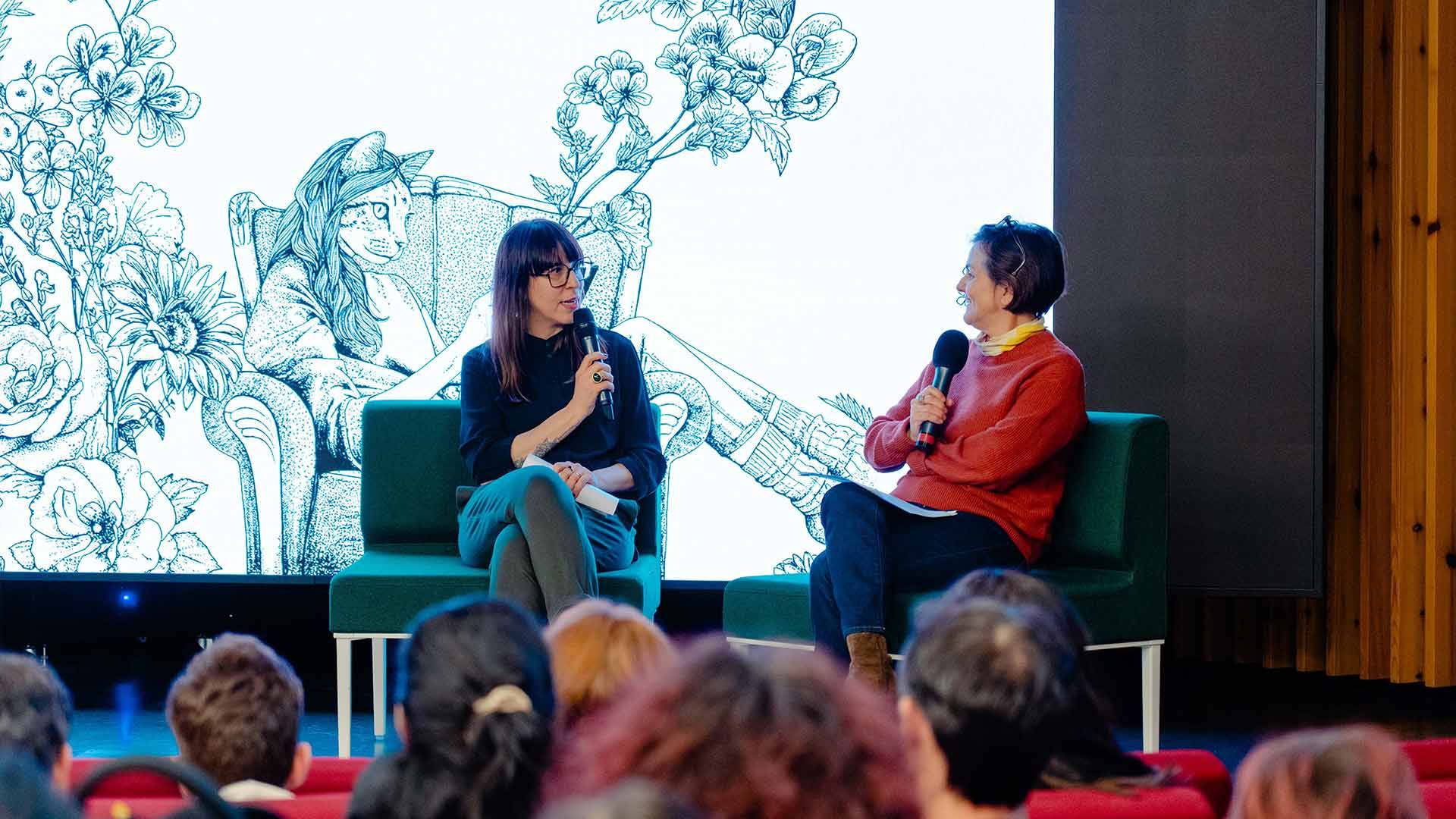 Dos mujeres conversan en un evento sobre un fondo artístico con ilustraciones de flores y un personaje sentado.