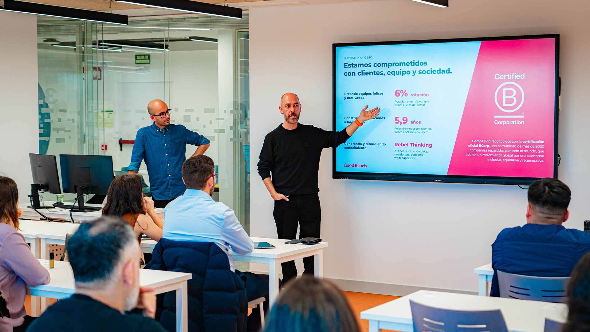 Una presentación en un aula donde dos ponentes hablan frente a un grupo de personas interesadas.