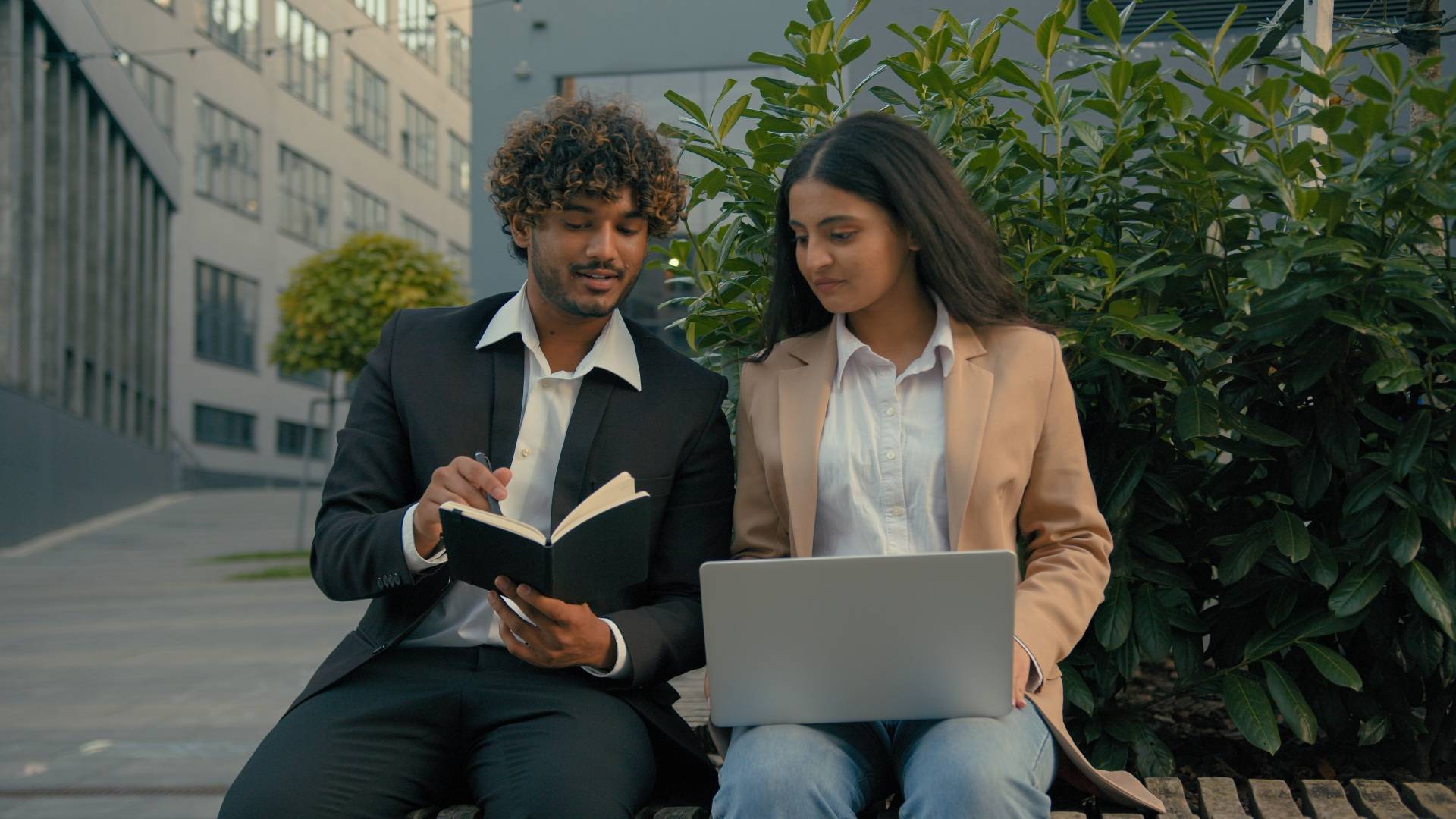 Dos personas conversando y trabajando al aire libre, una con un cuaderno y la otra con una computadora portátil.