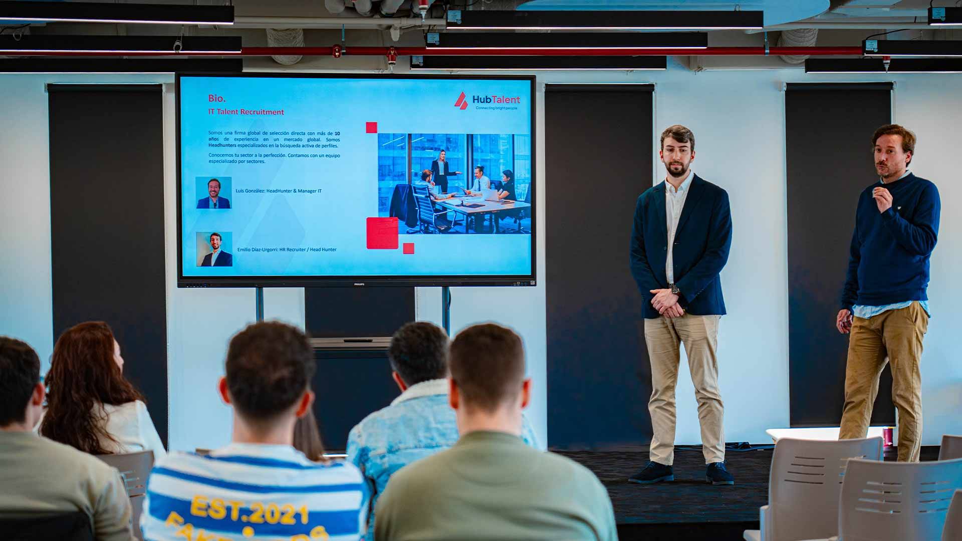 Dos presentadores están exponiendo en una sala frente a una audiencia mientras se proyecta una diapositiva en una pantalla.