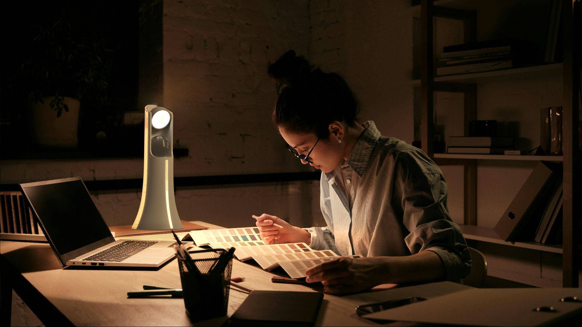 Una mujer está revisando un libro de muestras de colores en una habitación oscura, iluminada por una lámpara y con un ordenador portátil sobre la mesa.